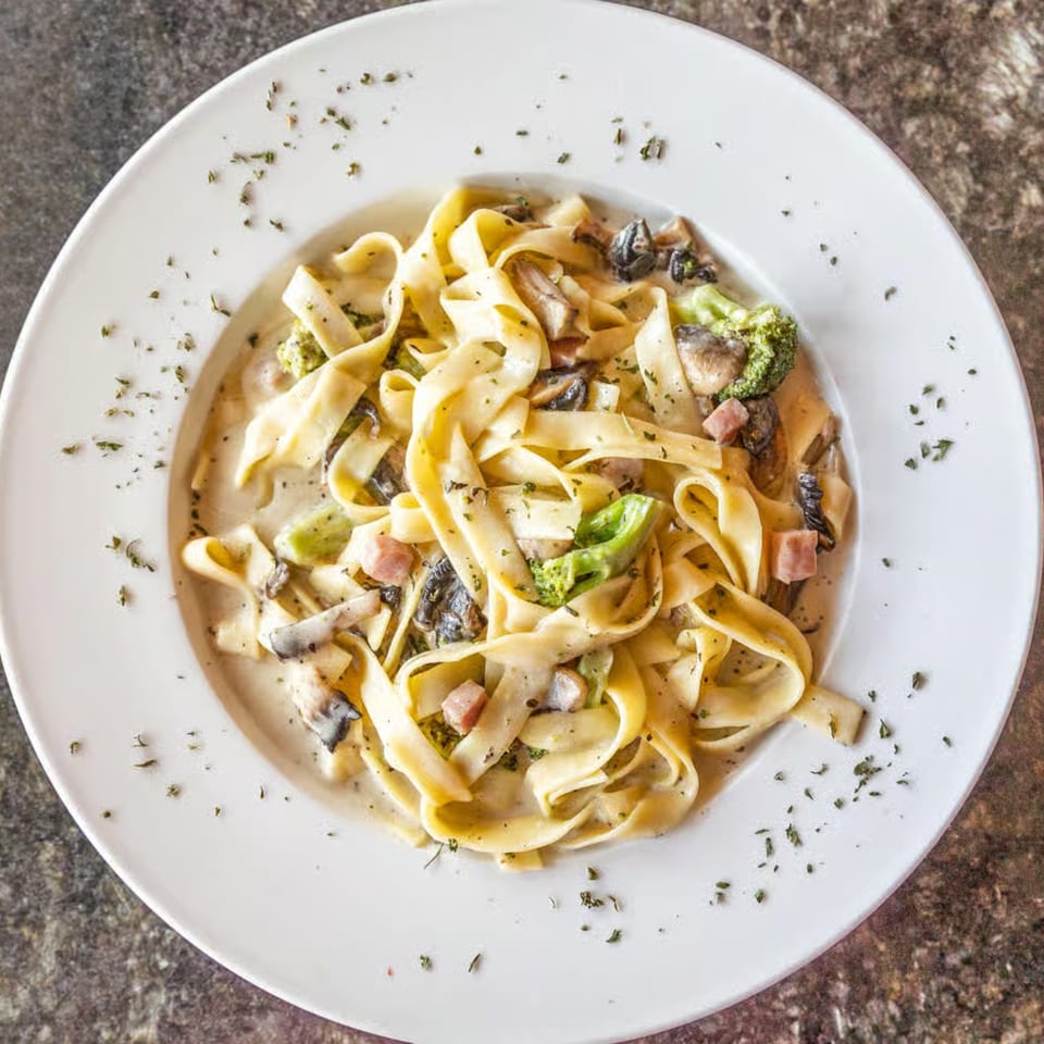 Creamy fettuccine pasta with mushrooms, broccoli, and ham in a white bowl, garnished with herbs.